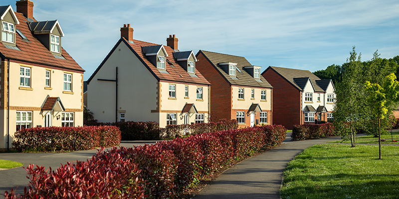 Image of a modern new-build housing estate with detached and semi-detached homes, landscaped paths, and red-leaved hedging — typical of developments like Kingsmere or Elmsbrook in Bicester.